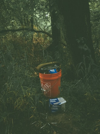 An orange bucket with a brand logo is placed on a patch of grass in a forested area. Inside the bucket are various tools or items, and next to it, there is a box labeled 'Fastenal.' The background features dense greenery and trees, with the lighting creating a shadowy and mysterious atmosphere.
