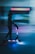 A person wearing a helmet rides an electric scooter with glowing blue lights in an urban environment at night. Motion blur conveys a sense of speed as they move past neon-lit cityscapes.