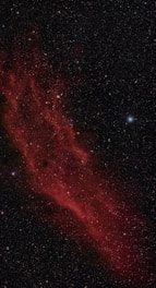 An expansive view of a nebula with a rich red color, surrounded by countless stars scattered across the dark background of space. The nebula stretches diagonally, with intricate feathery patterns and variations in luminosity, giving a sense of depth and texture.