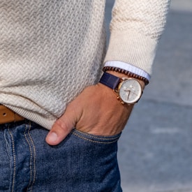 A person wearing a cream-colored knitted sweater and blue jeans has their hand in a pocket. On their wrist is a classic wristwatch with a leather strap and a bracelet made of brown beads. The watch has a white face and gold detailing.