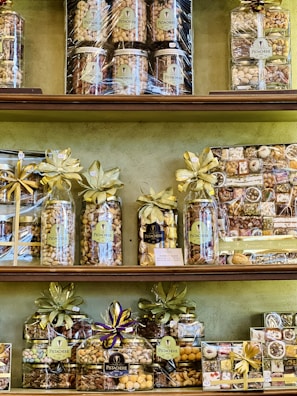 A variety of neatly arranged nut gift packages are displayed on shelves. Each package is wrapped with golden and purple ribbons and labeled with 'Pistacherie'. Different types of nuts and nut mixes can be seen, including pistachios.