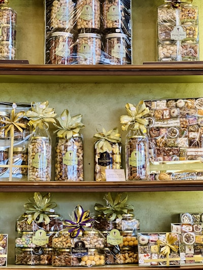 A variety of neatly arranged nut gift packages are displayed on shelves. Each package is wrapped with golden and purple ribbons and labeled with 'Pistacherie'. Different types of nuts and nut mixes can be seen, including pistachios.