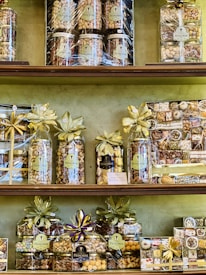 A variety of neatly arranged nut gift packages are displayed on shelves. Each package is wrapped with golden and purple ribbons and labeled with 'Pistacherie'. Different types of nuts and nut mixes can be seen, including pistachios.