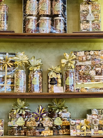 A variety of neatly arranged nut gift packages are displayed on shelves. Each package is wrapped with golden and purple ribbons and labeled with 'Pistacherie'. Different types of nuts and nut mixes can be seen, including pistachios.