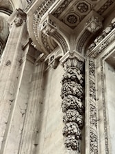 Intricate architectural details featuring ornate carvings and embellishments on stone columns and archways. The design includes floral and geometric patterns with remnants of historical artistry. Stone surfaces display weathering and patina, adding to the antique ambiance.