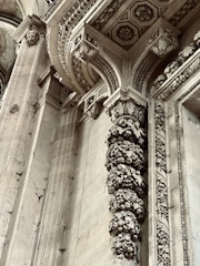 Intricate architectural details featuring ornate carvings and embellishments on stone columns and archways. The design includes floral and geometric patterns with remnants of historical artistry. Stone surfaces display weathering and patina, adding to the antique ambiance.