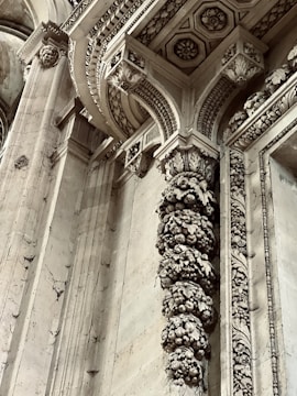 Intricate architectural details featuring ornate carvings and embellishments on stone columns and archways. The design includes floral and geometric patterns with remnants of historical artistry. Stone surfaces display weathering and patina, adding to the antique ambiance.