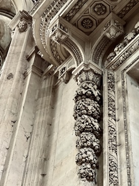 Intricate architectural details featuring ornate carvings and embellishments on stone columns and archways. The design includes floral and geometric patterns with remnants of historical artistry. Stone surfaces display weathering and patina, adding to the antique ambiance.