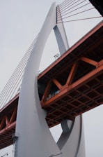 An architectural structure featuring a large, arch-like support made of concrete and orange metal sections. The bridge’s design includes tension cables supporting the roadbed and has a modern, industrial appearance.