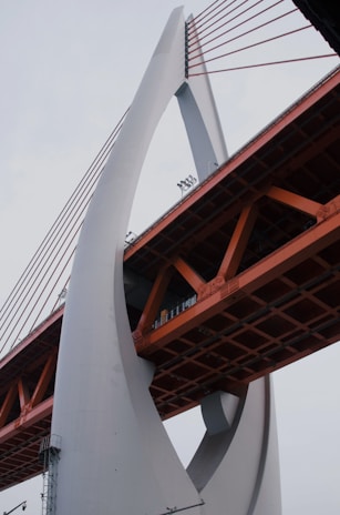 An architectural structure featuring a large, arch-like support made of concrete and orange metal sections. The bridge’s design includes tension cables supporting the roadbed and has a modern, industrial appearance.