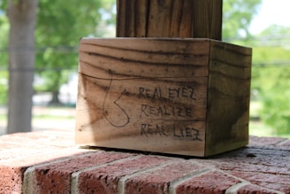 A wooden post is secured onto a brick surface. The wood has a rough, weathered texture, and there is black graffiti on it that reads 'REALEYEZ REALIZE REAL LIEZ'. The background is blurred, showing greenery and trees, indicating an outdoor setting.
