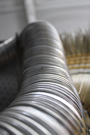 A tightly coiled metallic spiral, possibly part of a tubing or hose, is captured with a shallow depth of field. The shiny, metallic surface reflects light, creating a series of highlights and shadows. The background appears blurry, suggesting an industrial or mechanical setting.