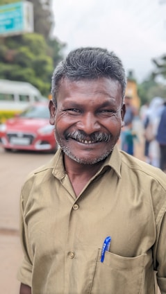 A middle-aged man with a broad smile wearing a beige shirt stands outdoors. He has short hair and a blue pen tucked into his shirt pocket. In the background, there are blurry images of a red car, greenery, and people walking.