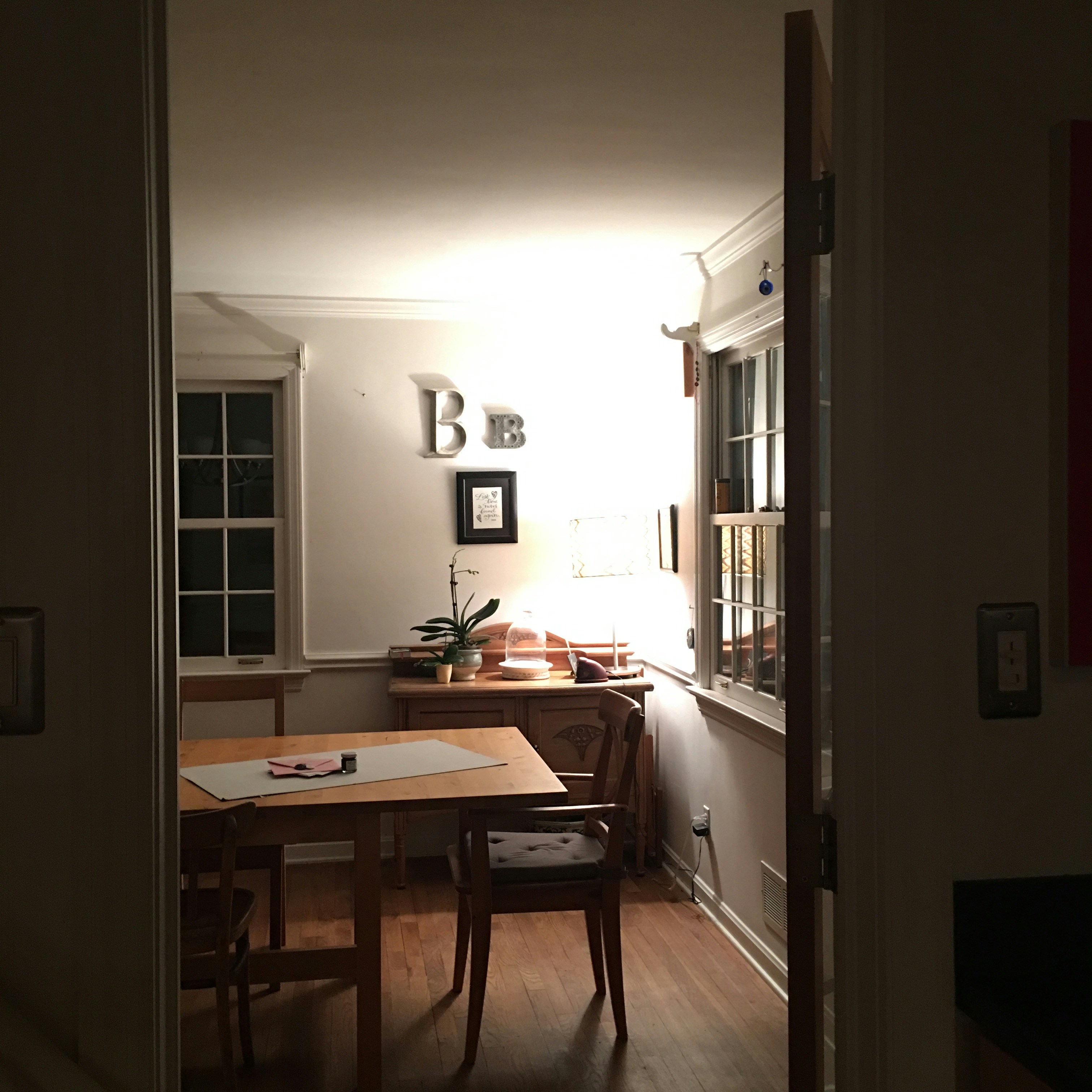 A cozy room features a wooden table with two chairs, and a small plant placed near a lamp on a sideboard. Wall art consisting of metallic letters 'B' is mounted above the sideboard. The room is warmly lit, contributing to a serene and inviting atmosphere.