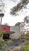 A building with a red and white facade, featuring a religious statue and the phrase 'COME TO ME' written on it. The entrance is labeled 'SHRINE OF THE INFANT JESUS' and is surrounded by greenery, including trees and plants. Overhead, branches with leaves partially obscure the sky.