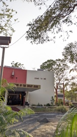 A building with a red and white facade, featuring a religious statue and the phrase 'COME TO ME' written on it. The entrance is labeled 'SHRINE OF THE INFANT JESUS' and is surrounded by greenery, including trees and plants. Overhead, branches with leaves partially obscure the sky.