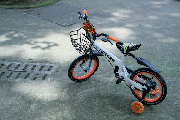 A small children's bicycle with training wheels is parked on a paved surface. It features a white frame with orange and black accents, including orange handle grips and wheel rims. A black basket is attached to the front, and the bike is decorated with a small bouquet of artificial flowers.