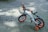 A small children's bicycle with training wheels is parked on a paved surface. It features a white frame with orange and black accents, including orange handle grips and wheel rims. A black basket is attached to the front, and the bike is decorated with a small bouquet of artificial flowers.