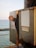 A metal electrical box with a red rotary switch mechanism is mounted on a pier. The surface of the box is rusted and weathered, with warning labels and a blank white label attached. The ocean is visible in the background, along with a section of metal grid railing.