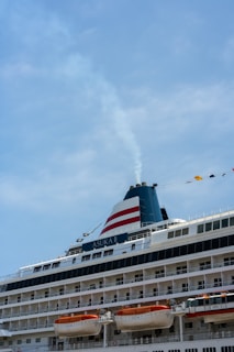 A large cruise ship with multiple decks and lifeboats visible, adorned with flags and a smokestack emitting a thin stream of smoke against a clear blue sky.