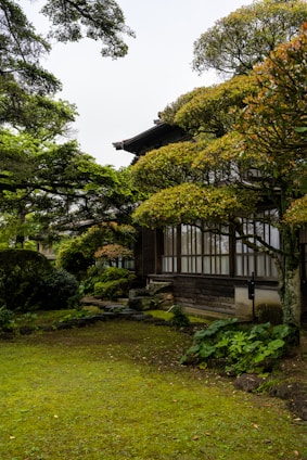 A traditional wooden building is surrounded by lush greenery. The architecture features large windows and dark wooden walls, blending harmoniously with the well-maintained garden. Trees and shrubs with dense foliage frame the building, and a small stone pathway leads through the mossy ground.