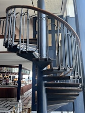A spiral staircase with a wooden handrail and dark metal balusters is prominently featured. It leads up through a modern, stylish interior that includes a bar area with various bottles and glassware, and seating visible in the background. The floor design is geometric, with tiles arranged in a pattern.