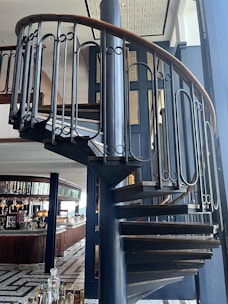 A spiral staircase with a wooden handrail and dark metal balusters is prominently featured. It leads up through a modern, stylish interior that includes a bar area with various bottles and glassware, and seating visible in the background. The floor design is geometric, with tiles arranged in a pattern.