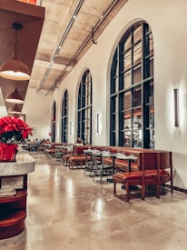 A modern, stylish restaurant with high ceilings and large arch windows. The seating area consists of red cushioned chairs and benches along tables, arranged neatly in rows. The lighting is warm, provided by multiple pendant lights hanging from the ceiling. There is a marble counter with a red flower arrangement near the left side of the image. A few people are seated in the background enjoying their meals.