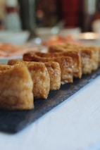 Several pieces of golden-brown fried tofu are neatly arranged in a row on a dark slate serving tray. The tofu has a crisp exterior and appears freshly cooked. The background is softly blurred, suggesting a dining setting with warm lighting.