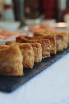 Several pieces of golden-brown fried tofu are neatly arranged in a row on a dark slate serving tray. The tofu has a crisp exterior and appears freshly cooked. The background is softly blurred, suggesting a dining setting with warm lighting.