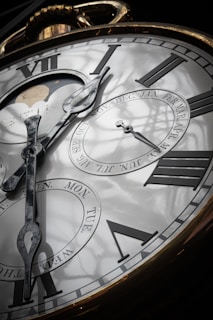 An ornate, close-up view of a large clock face with Roman numerals. The clock hands point to various times and there are additional dials showing days of the week and months. A moon phase display is visible, and the reflective surface reveals shadows of a window frame.