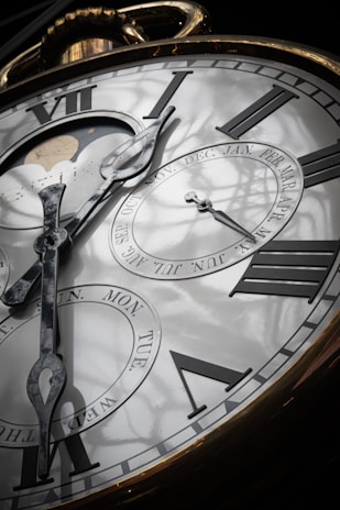 An ornate, close-up view of a large clock face with Roman numerals. The clock hands point to various times and there are additional dials showing days of the week and months. A moon phase display is visible, and the reflective surface reveals shadows of a window frame.