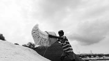 A person is engaged in pouring a large quantity of white substance, possibly salt, from a container onto a large pile. The scene is set outdoors under a cloudy sky, with various shapes and textures adding depth to the landscape.