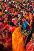 A vibrant gathering of people, predominantly women, dressed in colorful traditional attire, participating in a cultural or religious event. Many are holding offerings or ceremonial items such as fruits and flowers. The crowd is dense, with individuals and groups engaged in various activities, creating an atmosphere of festivity and devotion.