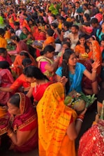 A vibrant gathering of people, predominantly women, dressed in colorful traditional attire, participating in a cultural or religious event. Many are holding offerings or ceremonial items such as fruits and flowers. The crowd is dense, with individuals and groups engaged in various activities, creating an atmosphere of festivity and devotion.
