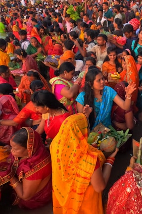A vibrant gathering of people, predominantly women, dressed in colorful traditional attire, participating in a cultural or religious event. Many are holding offerings or ceremonial items such as fruits and flowers. The crowd is dense, with individuals and groups engaged in various activities, creating an atmosphere of festivity and devotion.