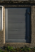A metallic louvered ventilation panel set within a roughly textured, pebble-dash wall. The panel is rectangular with horizontal slats and secured by metal hinges on the right side. Green plants grow at the base against the concrete surface.
