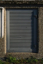 A metallic louvered ventilation panel set within a roughly textured, pebble-dash wall. The panel is rectangular with horizontal slats and secured by metal hinges on the right side. Green plants grow at the base against the concrete surface.