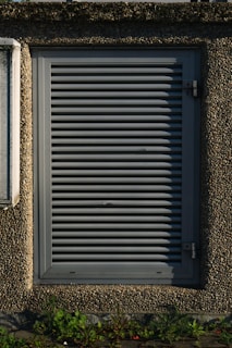 A metallic louvered ventilation panel set within a roughly textured, pebble-dash wall. The panel is rectangular with horizontal slats and secured by metal hinges on the right side. Green plants grow at the base against the concrete surface.