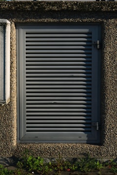 A metallic louvered ventilation panel set within a roughly textured, pebble-dash wall. The panel is rectangular with horizontal slats and secured by metal hinges on the right side. Green plants grow at the base against the concrete surface.