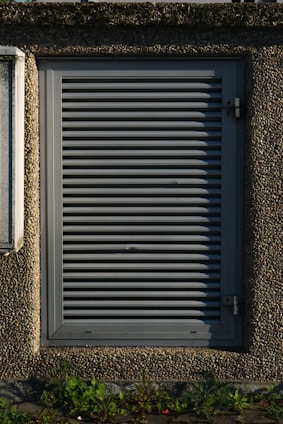 A metallic louvered ventilation panel set within a roughly textured, pebble-dash wall. The panel is rectangular with horizontal slats and secured by metal hinges on the right side. Green plants grow at the base against the concrete surface.