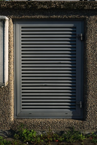 A metallic louvered ventilation panel set within a roughly textured, pebble-dash wall. The panel is rectangular with horizontal slats and secured by metal hinges on the right side. Green plants grow at the base against the concrete surface.