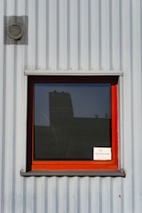 A corrugated metal wall with a red-framed window and a small sign displayed in the lower right corner of the window. The wall is industrial-looking, featuring a small vent near the upper left corner, and the window reflects part of a building.