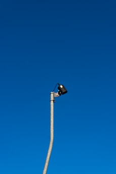 A tall streetlight pole extends into a clear blue sky, with a security camera mounted near the top.