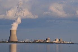 A nuclear power plant is situated by a body of water with a large cooling tower emitting steam. The sky is filled with scattered clouds, and the plant is reflected in the calm water surface.