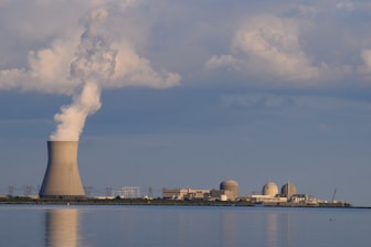 A nuclear power plant is situated by a body of water with a large cooling tower emitting steam. The sky is filled with scattered clouds, and the plant is reflected in the calm water surface.