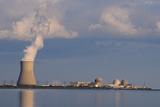 A nuclear power plant is situated by a body of water with a large cooling tower emitting steam. The sky is filled with scattered clouds, and the plant is reflected in the calm water surface.