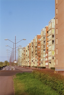 A row of modern apartment buildings extends into the distance alongside a road, with streetlights lining the pavement. The buildings feature a mix of brick and glass exteriors, and there is a neatly trimmed grassy area with some small shrubs in the foreground. The sky is clear and blue, suggesting a bright and sunny day.