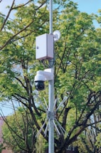 A surveillance camera mounted on a pole with a protective box and metal spikes below. The background features lush green trees and a clear blue sky, suggesting a peaceful outdoor setting.
