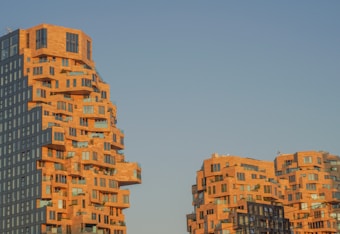 Unique architectural buildings with a modern design, featuring staggered and irregular shapes. The structures have a mix of glass and brick exteriors and are set against a clear blue sky. The sunlight casts a warm, golden hue over the buildings, highlighting their geometric patterns.